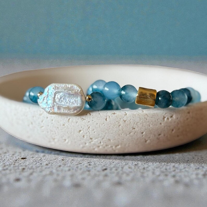 A beaded bracelet with light blue translucent beads, a textured white shell charm, and a small gold spacer, displayed on a white ceramic dish.