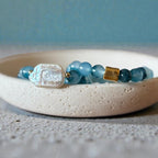 A beaded bracelet with light blue translucent beads, a textured white shell charm, and a small gold spacer, displayed on a white ceramic dish.
