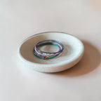 A stack of three beaded wire bracelet with blue, purple, and teal beads sits in a small white ceramic dish on a warm beige background.