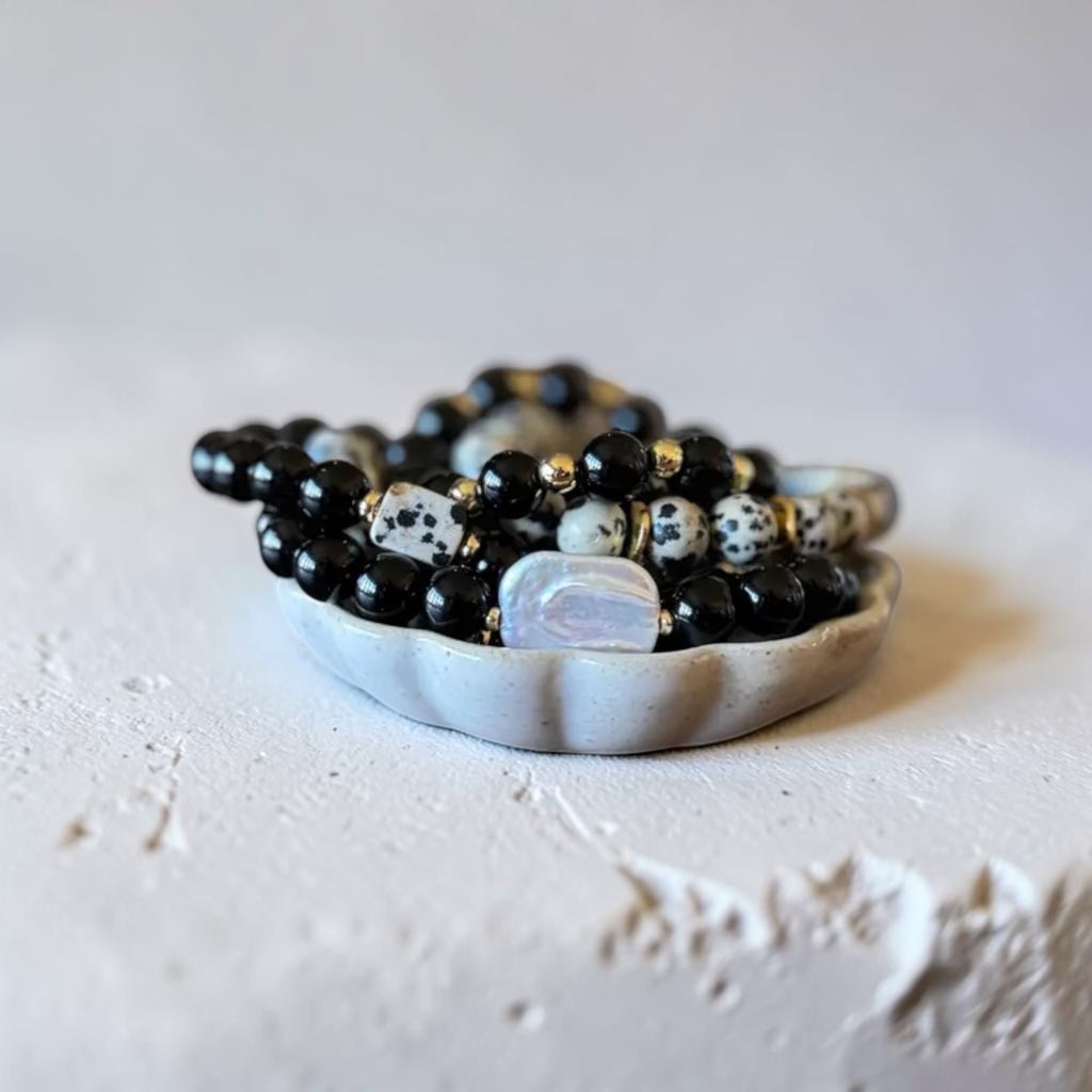 A beaded bracelet or necklace with glossy black beads, white-and-black spotted accent beads, and a large iridescent white oval charm, displayed in a small white dish on a light textured surface.
