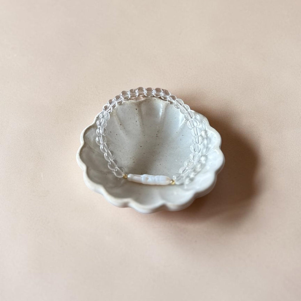 A delicate clear beaded bracelet with a small white pearl accent lies inside a scalloped, speckled white ceramic dish on a solid beige background.