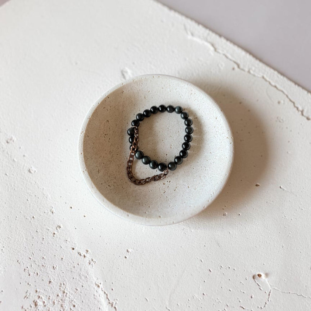 A beaded bracelet with dark round beads and a small bronze-toned metal loop, displayed inside a small round ceramic dish on a textured off-white surface.