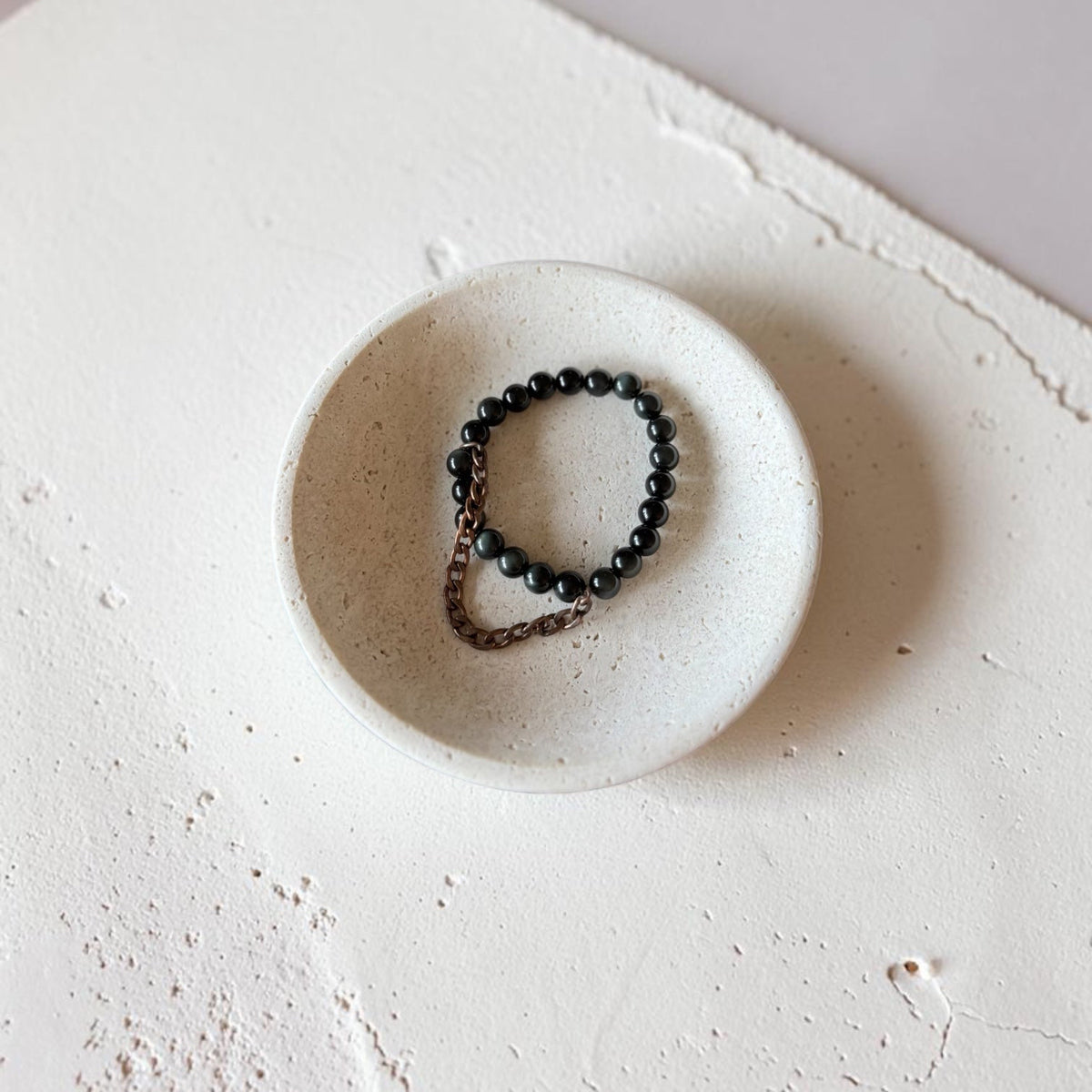 A beaded bracelet with dark round beads and a small bronze-toned metal loop, displayed inside a small round ceramic dish on a textured off-white surface.