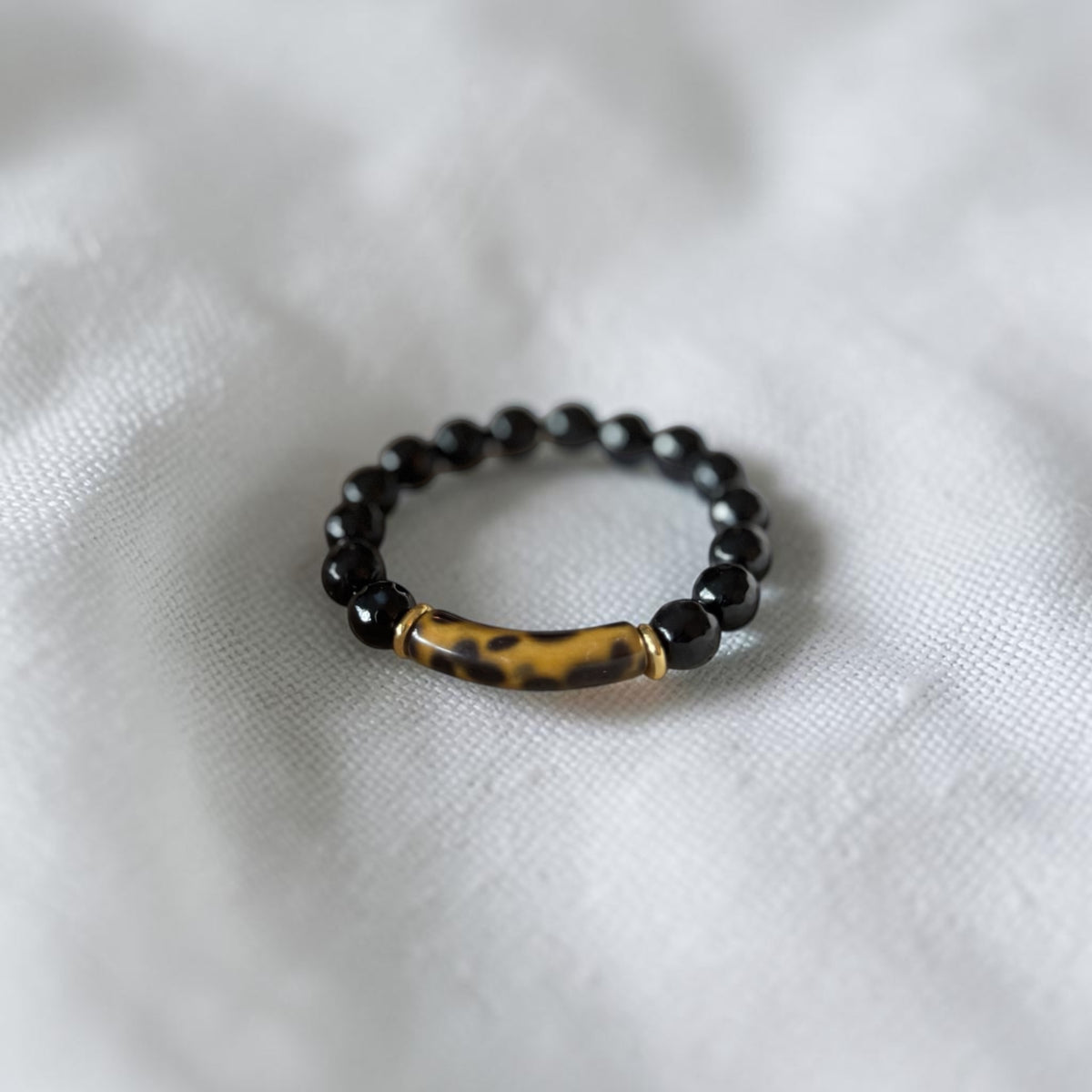 A beaded ring with black round beads and a central tortoiseshell barrel bead, displayed on a soft white textured fabric background.