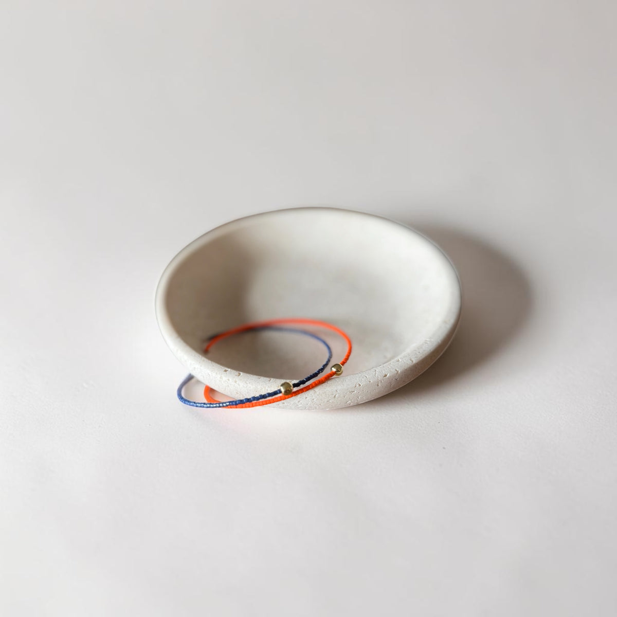 Two delicate wire bracelets, one orange and one blue, each with a small gold bead, displayed inside a small white ceramic dish on a light gray background.