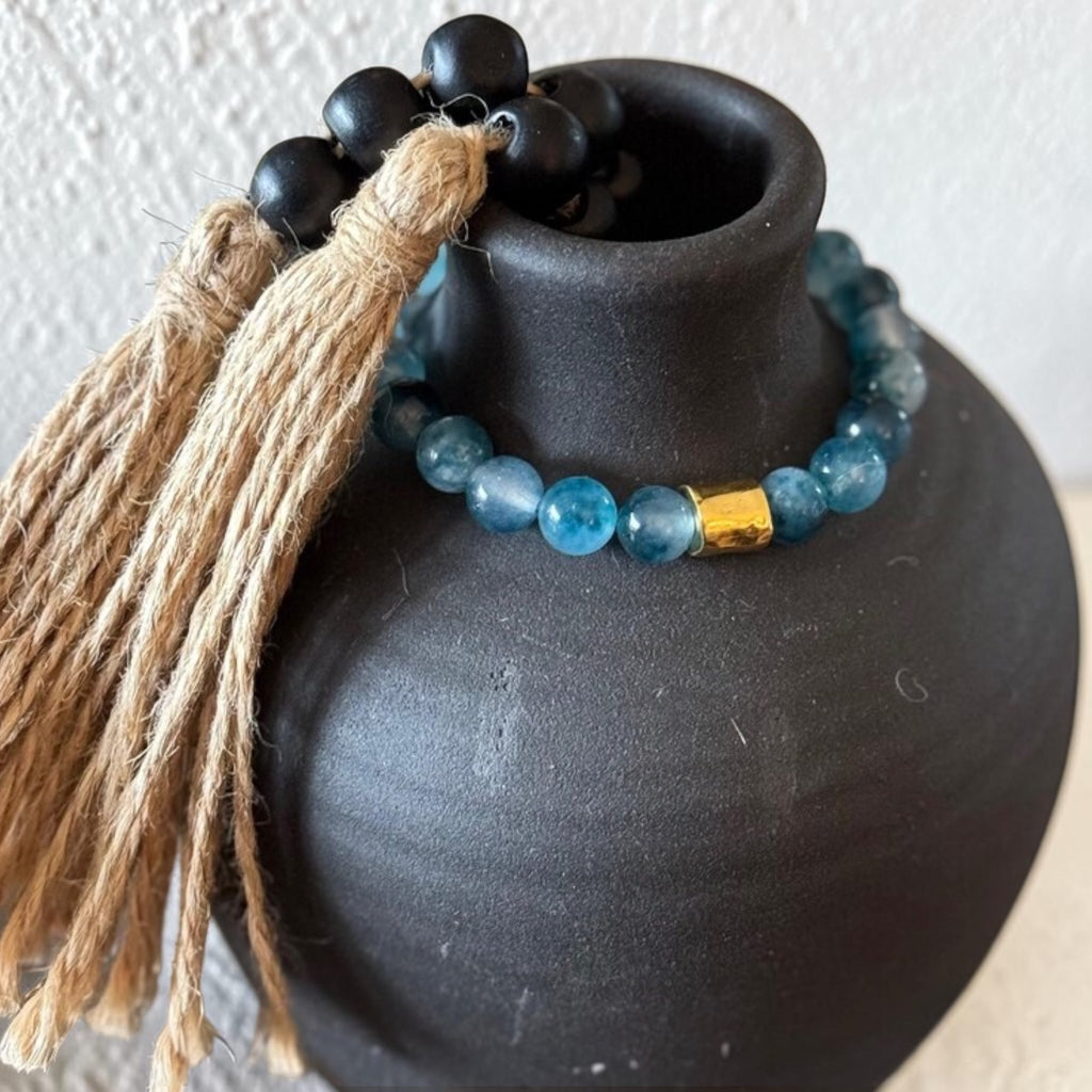 A beaded necklace with light blue round beads, a small gold spacer, and a black beaded tassel with tan jute wrapping, displayed on a matte black ceramic vase against a light textured wall.