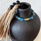 A beaded necklace with light blue round beads, a small gold spacer, and a black beaded tassel with tan jute wrapping, displayed on a matte black ceramic vase against a light textured wall.