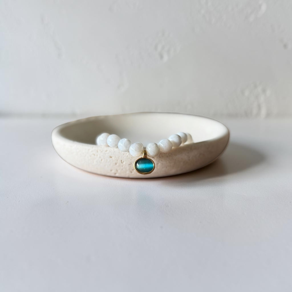 a white swirl bead bracelet with a gold charm that dangles. Inside the charm is a stone. It is sitting in a ceramic dish.