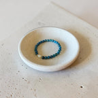 A beaded stretch bracelet with blue and teal beads and a small white pearl accent, displayed inside a small round ceramic dish on a textured beige surface.
