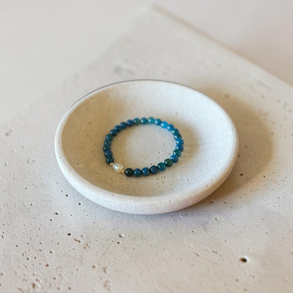 A beaded stretch bracelet with blue and teal beads and a small white pearl accent, displayed inside a small round ceramic dish on a textured beige surface.