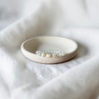 A clear-toned beaded bracelet with small white pearls and gold spacer beads rests on a shallow, round beige ceramic dish, set against a soft, draped white fabric background.