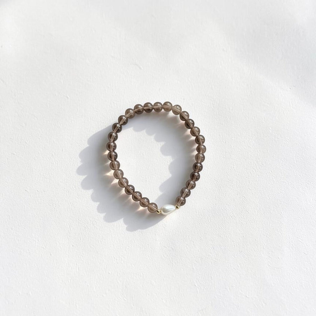 A beaded stretch bracelet made of small, translucent smoky brown beads with a single white pearl accent, photographed on a white textured surface with soft shadows.