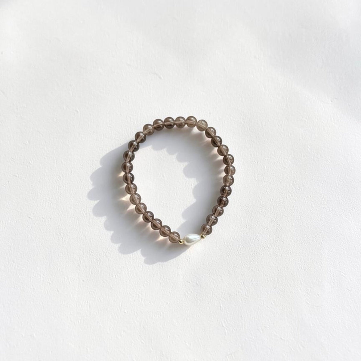 A beaded stretch bracelet made of small, translucent smoky brown beads with a single white pearl accent, photographed on a white textured surface with soft shadows.