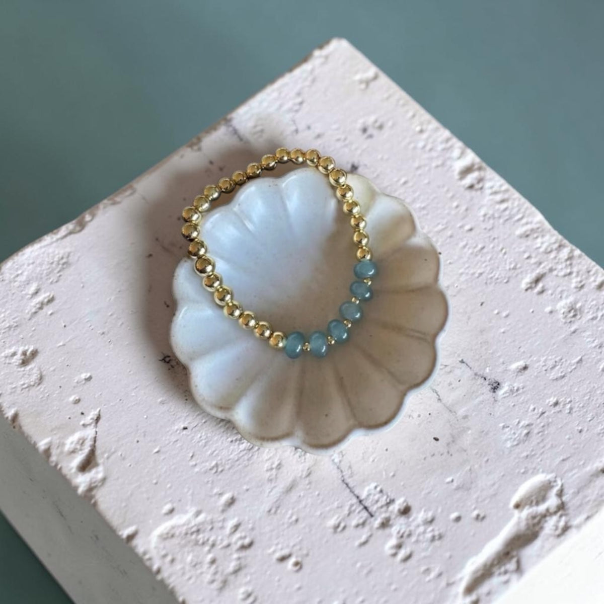 A delicate beaded bracelet with a gold-tone beaded band and a short section of light blue beads, displayed on a white scalloped shell-shaped stand.