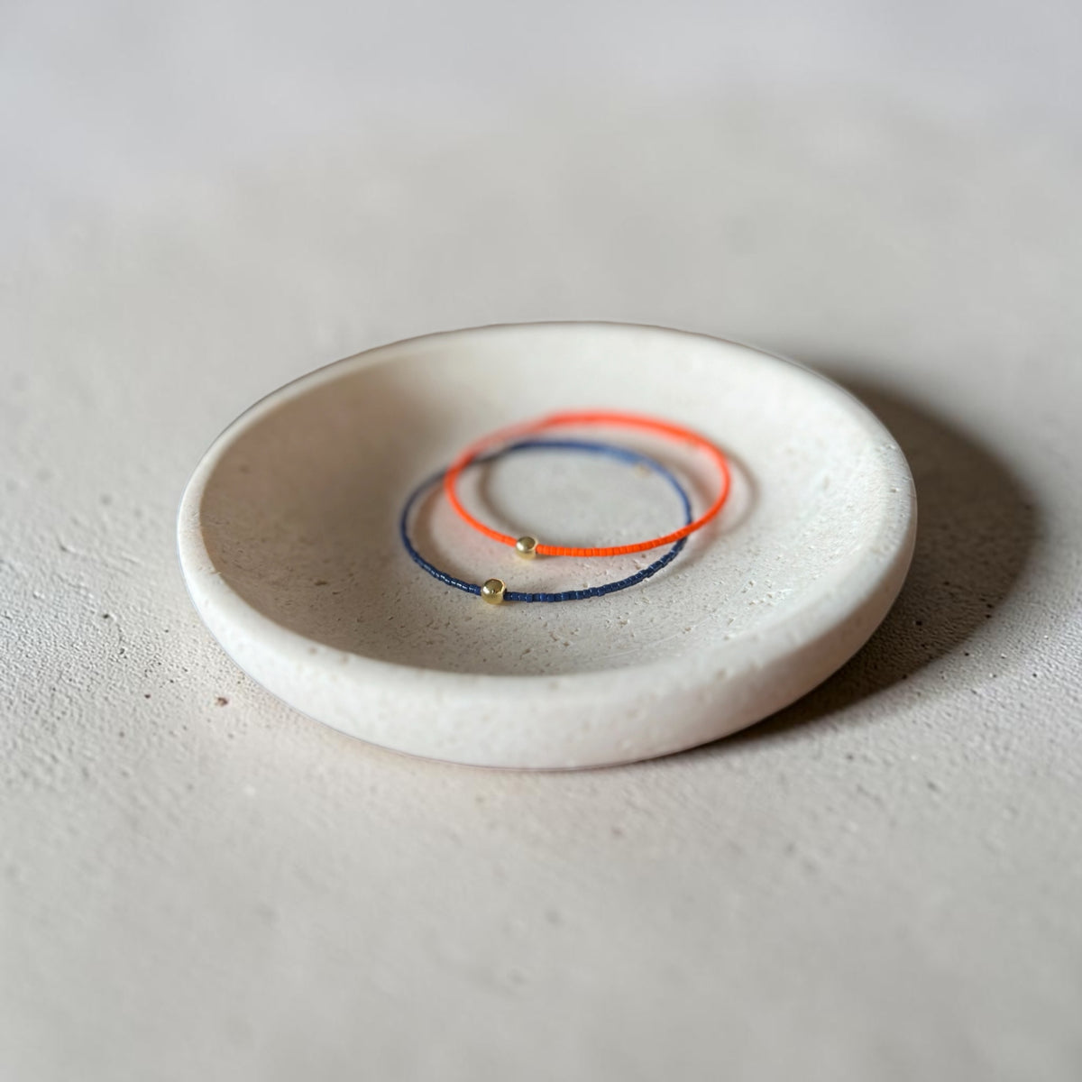 Two delicate cord bracelets, one orange and one blue, each with a small gold bead, displayed in a small round white ceramic dish on a textured light gray surface.