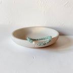 A beaded bracelet with light green round beads and a small white pearl accent, displayed on a small beige ceramic dish against a textured off-white background.