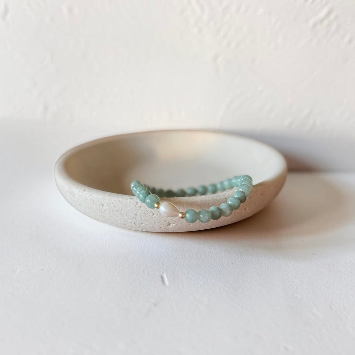 A beaded bracelet with light green round beads and a small white pearl accent, displayed on a small beige ceramic dish against a textured off-white background.