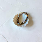 A beaded stretch bracelet made of polished tiger's eye beads in brown and black, displayed on a small white scalloped dish against a textured white background.