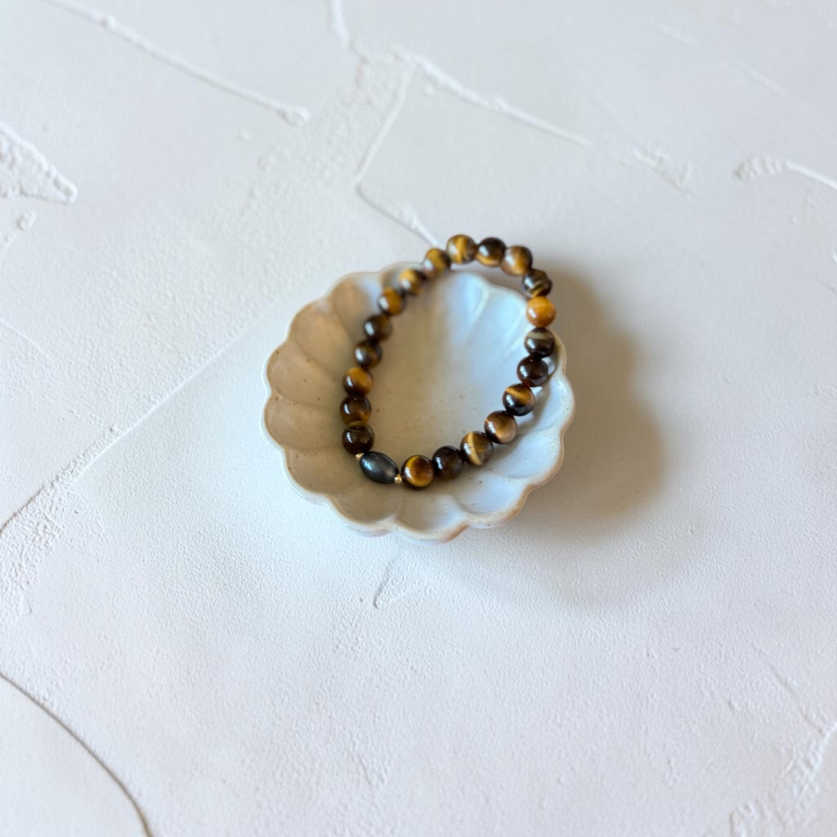 A beaded stretch bracelet made of polished tiger's eye beads in brown and black, displayed on a small white scalloped dish against a textured white background.