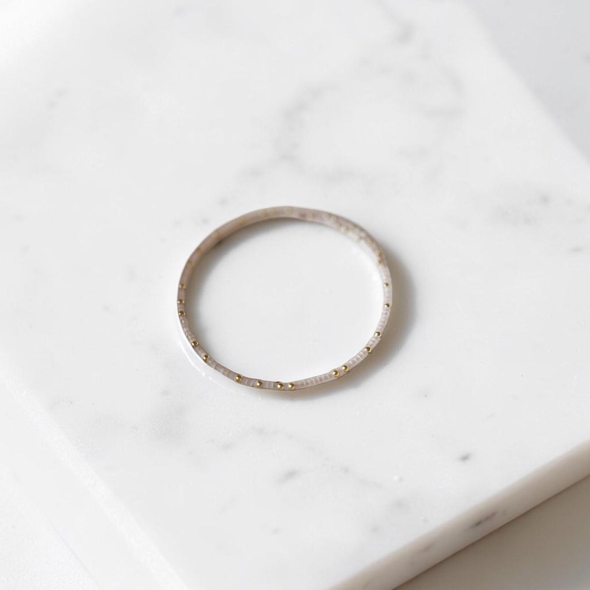 A thin seed bead wire bracelet in cream opal color with tiny gold dot accents along the band, resting on a white marble surface.