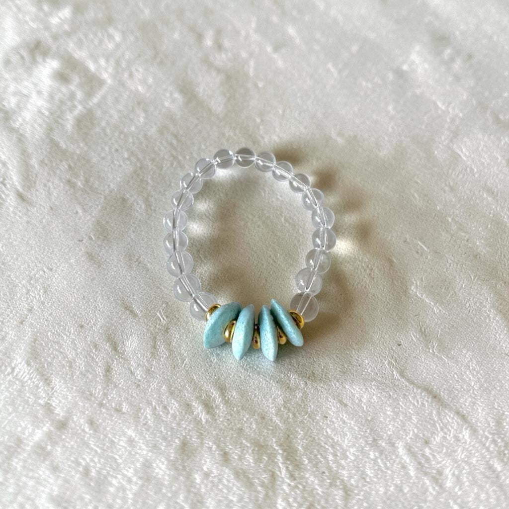 A beaded stretch ring featuring a clear, translucent bead band with five light blue enamel petal charms and small gold spacer rings, displayed on a textured white fabric background.
