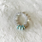A beaded stretch ring featuring a clear, translucent bead band with five light blue enamel petal charms and small gold spacer rings, displayed on a textured white fabric background.