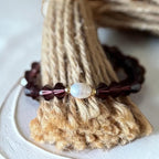 A beaded bracelet with dark burgundy faceted beads, a central white iridescent bead, and small gold spacer beads, displayed on a textured beige jute stand.