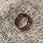 A beaded stretch bracelet featuring dark red beads and a small gold-toned triangular charm, displayed on a textured beige burlap fabric background.