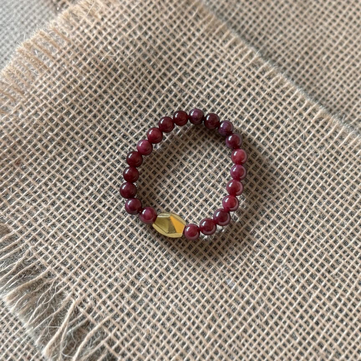 A beaded stretch bracelet featuring dark red beads and a small gold-toned triangular charm, displayed on a textured beige burlap fabric background.