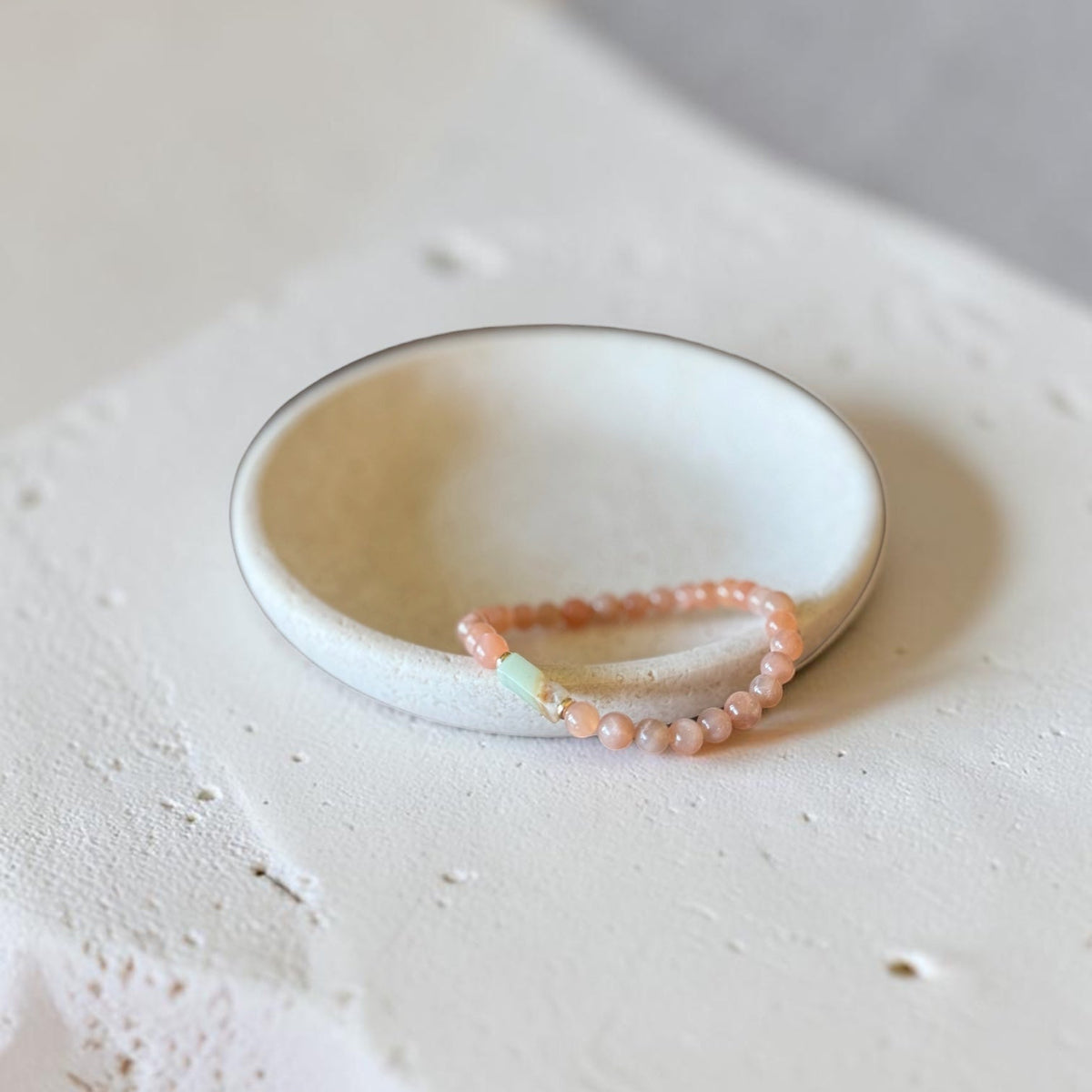 A delicate peach beaded bracelet with a small mint green bead accent, displayed inside a white ceramic dish on a textured beige surface.