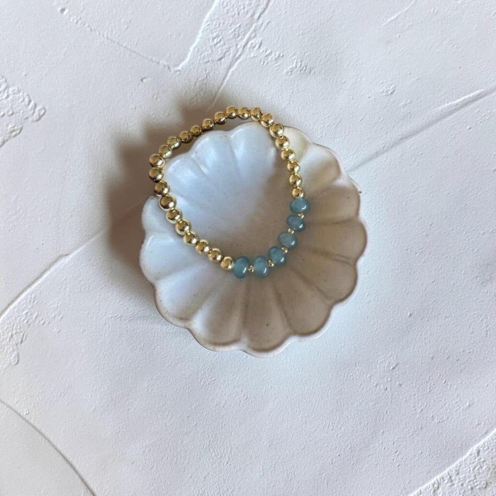 A beaded stretch bracelet with gold-toned beads and light blue-green beads, displayed on a white scalloped dish against a textured off-white background.