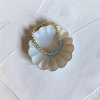 A beaded stretch bracelet with gold-toned beads and light blue-green beads, displayed on a white scalloped dish against a textured off-white background.