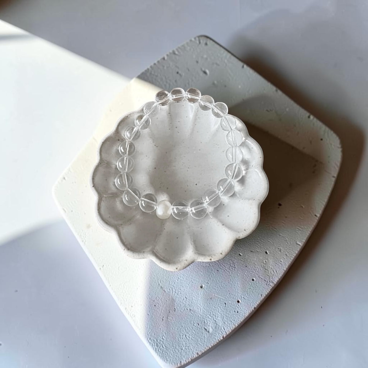 A clear crystal beaded bracelet with a central pearl-like bead, displayed on a small white scalloped ceramic dish over a textured white square surface.