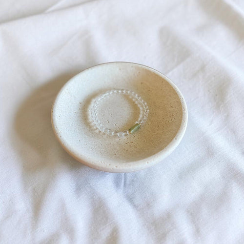 A small round ceramic dish with a speckled cream and light brown glaze, containing a small bracelet of white beads, set on a wrinkled white fabric background.