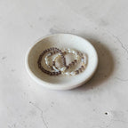 Three beaded bracelets in brown, white, and gray tones arranged in a small round white ceramic dish on a textured light gray surface.