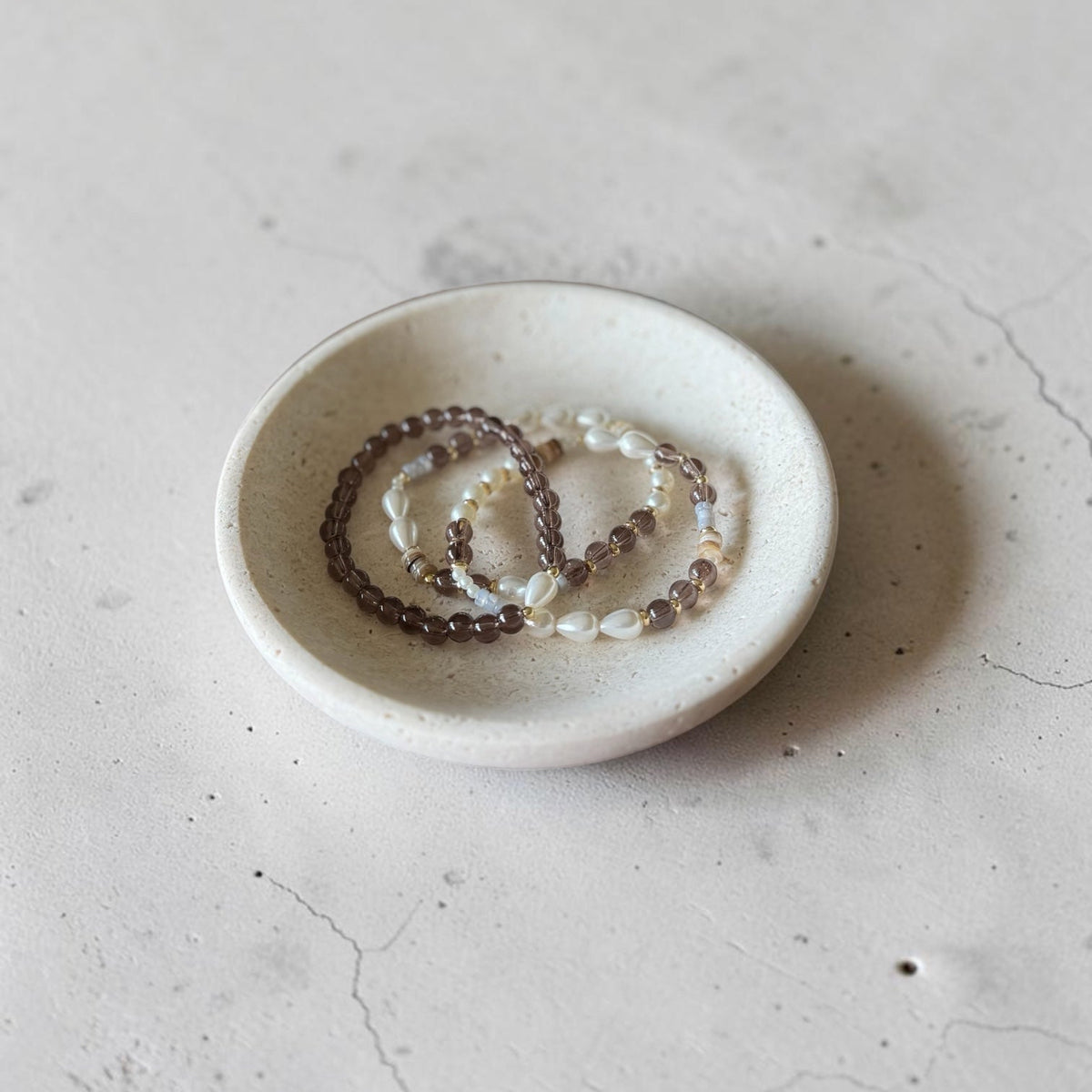 Three beaded bracelets in brown, white, and gray tones arranged in a small round white ceramic dish on a textured light gray surface.