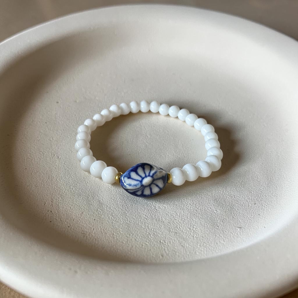 A beaded stretch bracelet with white round beads and a central blue and white floral disc charm, displayed on a textured beige ceramic dish.