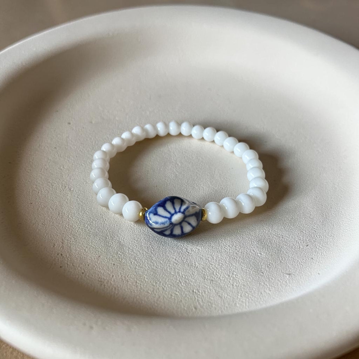 A beaded stretch bracelet with white round beads and a central blue and white floral disc charm, displayed on a textured beige ceramic dish.