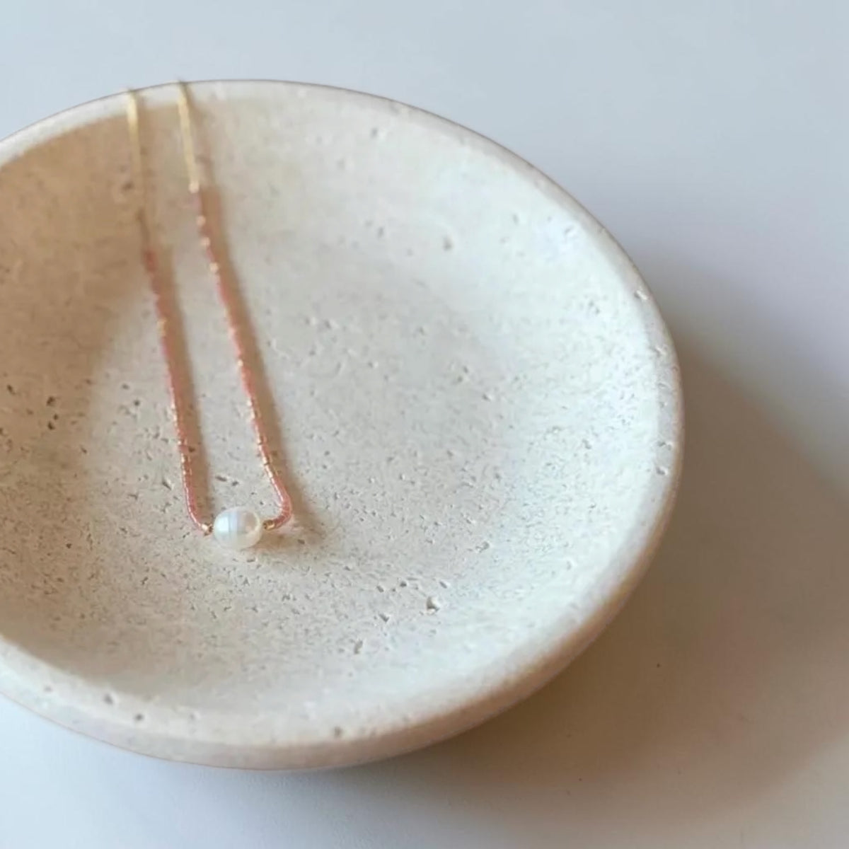A delicate necklace with a small white pearl and fine gold chain, displayed inside a round, speckled off-white ceramic dish on a light gray background.