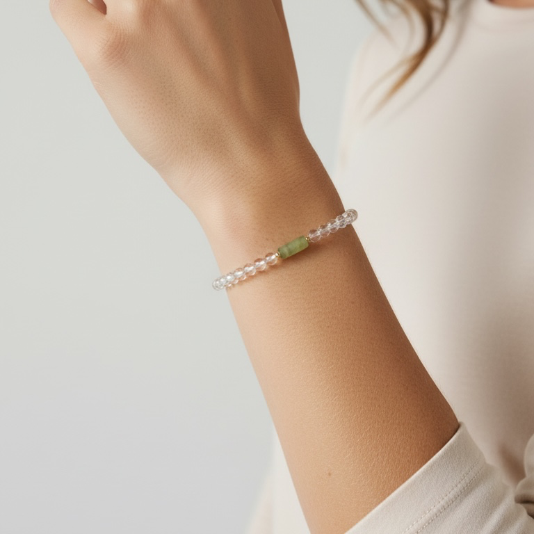 A close-up of a person's wrist wearing a delicate beaded bracelet with clear faceted beads and a small light green stone accent, against a soft light gray background.