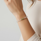 A close-up of a person's wrist wearing a delicate beaded bracelet with clear faceted beads and a small light green stone accent, against a soft light gray background.