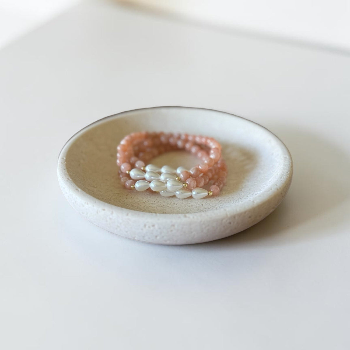 Three beaded stretch bracelets, including two peach-pink beaded ones and one white pearl strand, displayed on a small round ceramic dish.