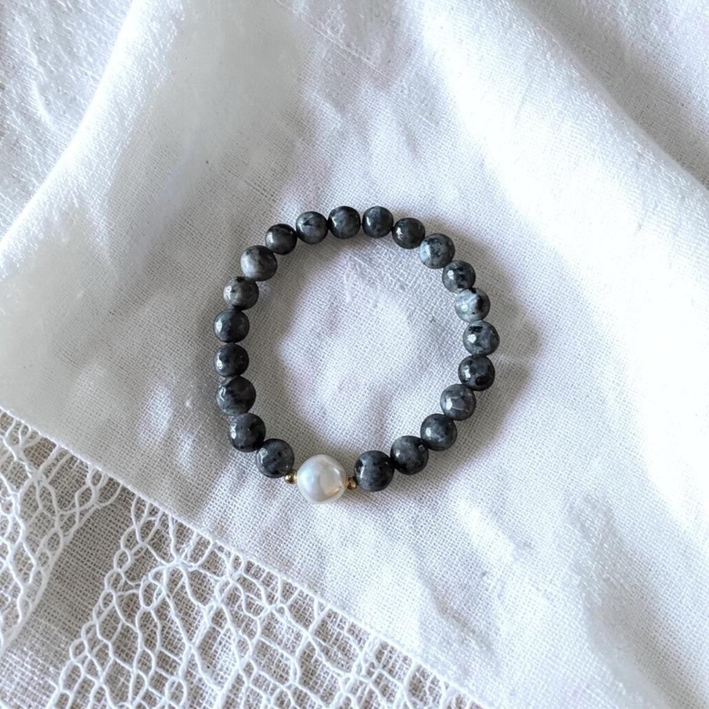 A beaded stretch bracelet with dark gray marbled beads and a single white pearl accent, displayed on a textured white fabric background.