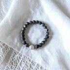 A beaded stretch bracelet with dark gray marbled beads and a single white pearl accent, displayed on a textured white fabric background.