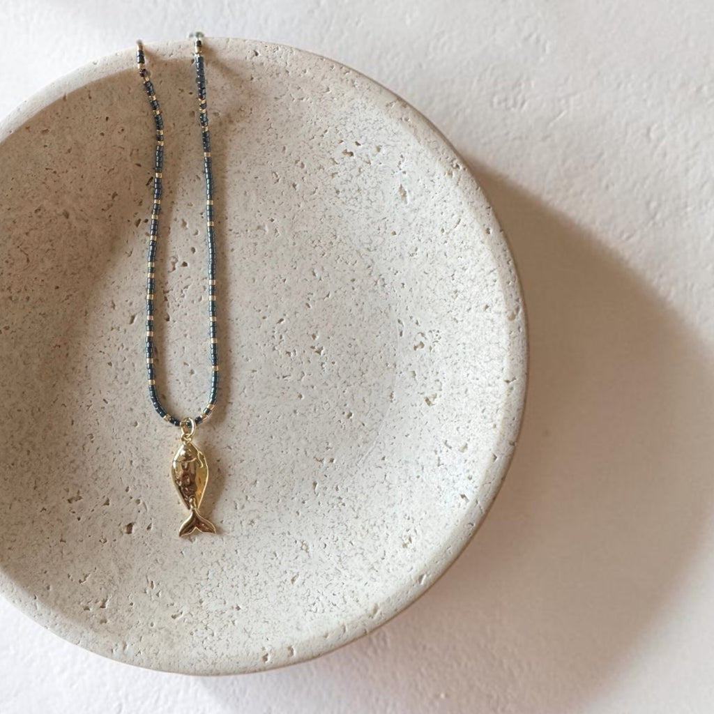 A delicate necklace with a small gold fish pendant and a blue beaded chain, displayed on a textured beige ceramic dish against a neutral off-white background.