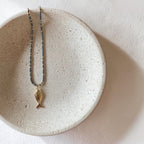 A delicate necklace with a small gold fish pendant and a blue beaded chain, displayed on a textured beige ceramic dish against a neutral off-white background.