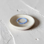 A delicate light blue beaded ring with a small white bead accent, displayed on a small round ceramic dish over a textured light gray surface.