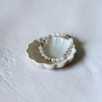 A delicate beaded bracelet with white pearls, light pink faceted beads, and small metallic spacer beads, displayed on a small white ceramic dish against a textured light gray background.