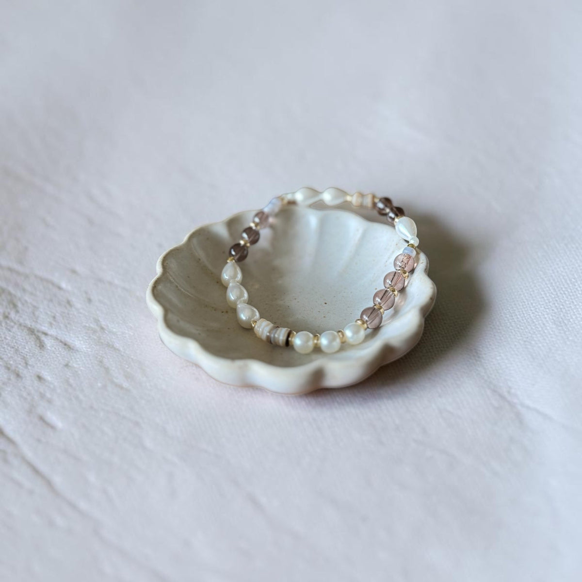 A delicate beaded bracelet with white pearls, light pink faceted beads, and small metallic spacer beads, displayed on a small white ceramic dish against a textured light gray background.
