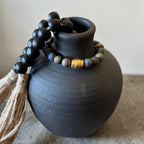 Black wooden prayer beads with a tassel and a small gold accent bead, draped over a matte black ceramic vase against a neutral background.